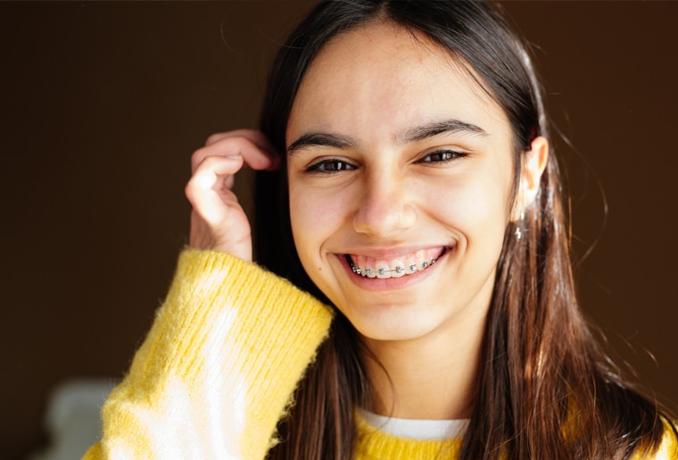 Smiling teenage girl in yellow sweater touching her hair against brown background.