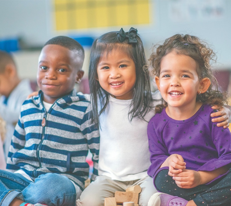 Three young children smiling together in a classroom setting with colorful background.