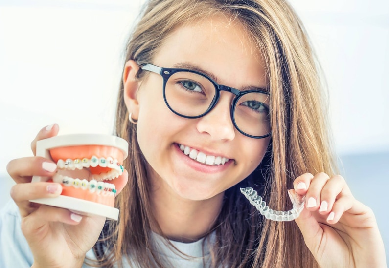 Young girl wearing glasses smiles while holding orthodontic model and dental aligners outdoors.