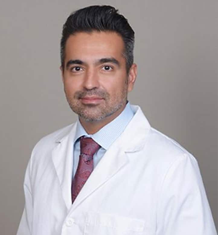 Professional headshot of male orthodontist in white coat and burgundy tie against gray background.