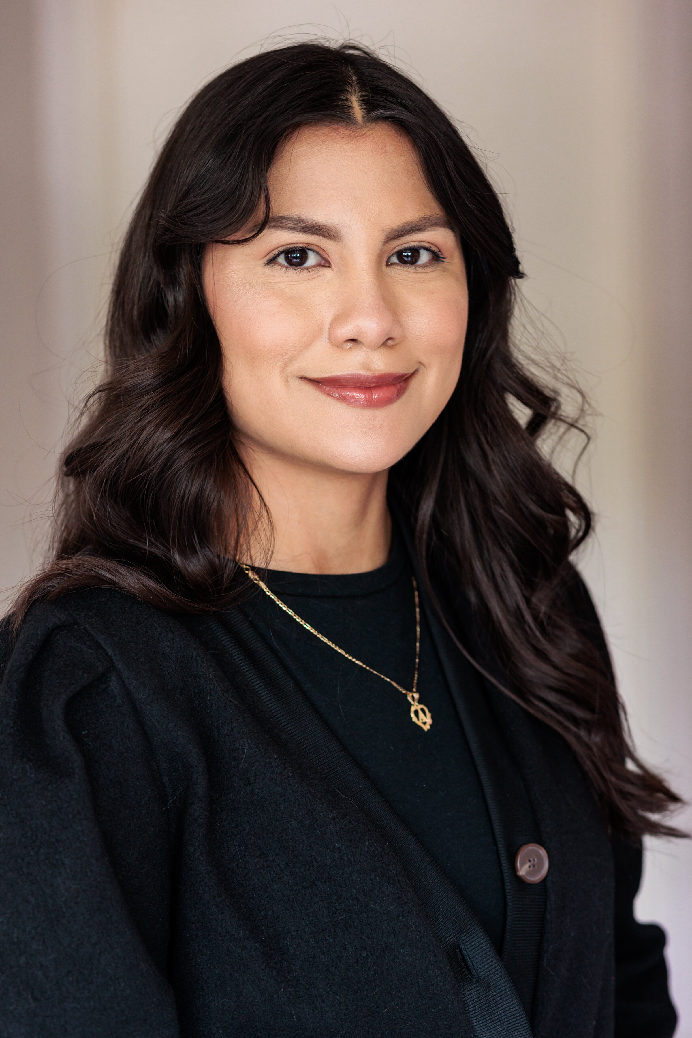 Professional headshot of woman with long dark hair wearing black blazer and gold necklace against neutral background.