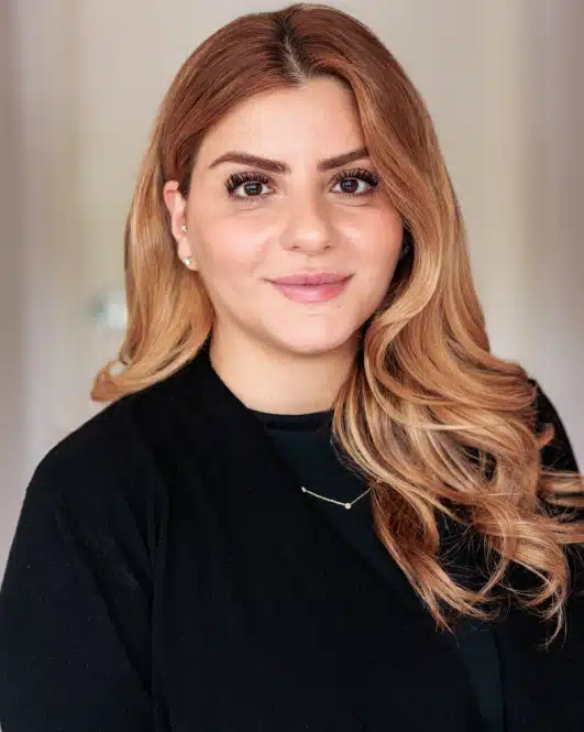 Professional headshot of a woman with auburn wavy hair wearing black, against neutral background.