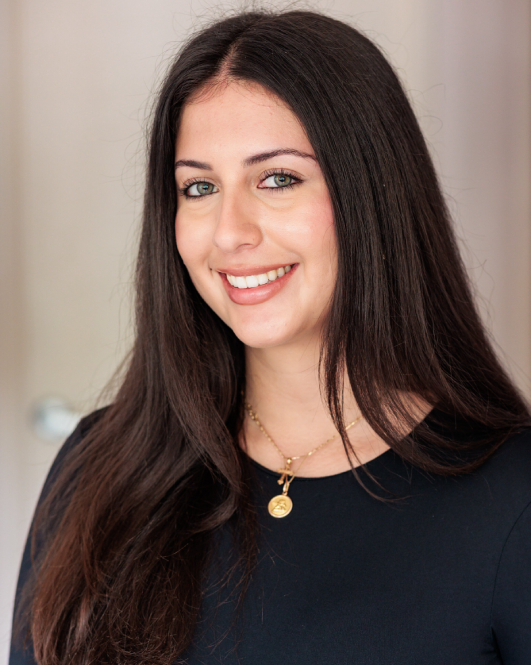 Professional headshot of a smiling woman with long dark hair wearing a black top and gold pendant necklace.