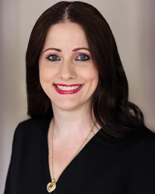 Professional headshot of woman with long dark hair, pink lipstick, wearing black blazer against neutral background.