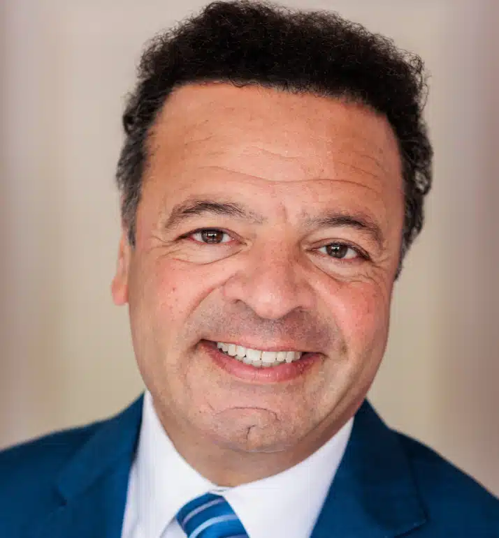 Professional headshot of smiling man in blue suit and tie against neutral background.