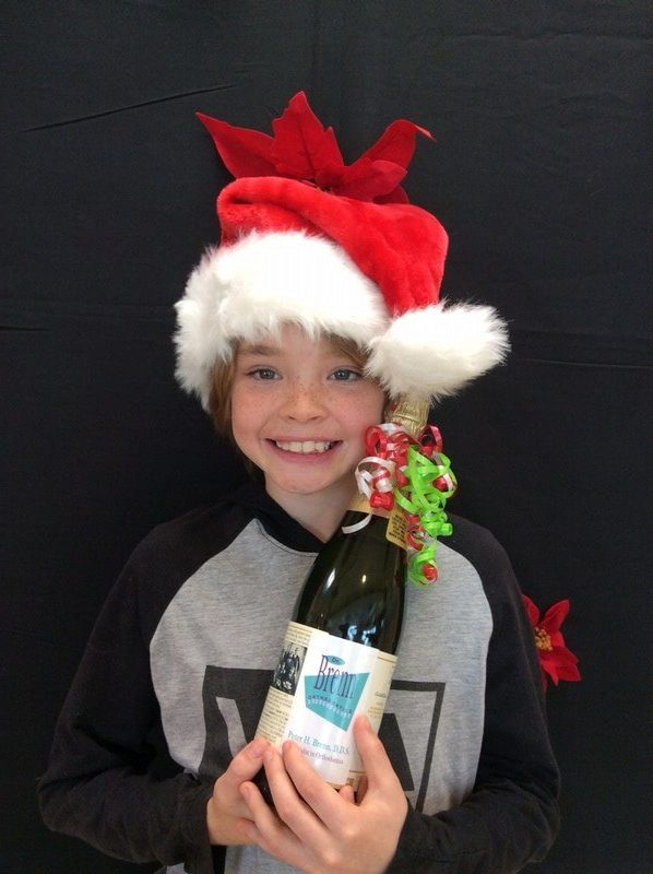 Young child wearing red Santa hat holding gift box against black background, smiling at camera.