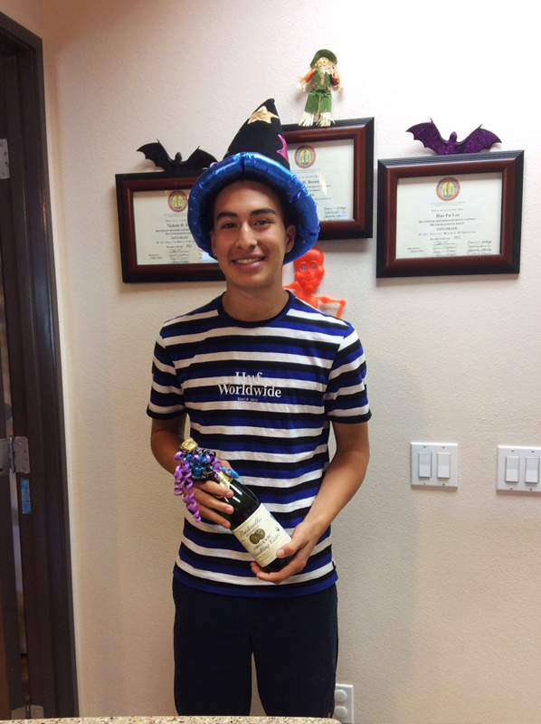 Young man in striped shirt wearing blue crown, holding award in office with framed certificates on wall.