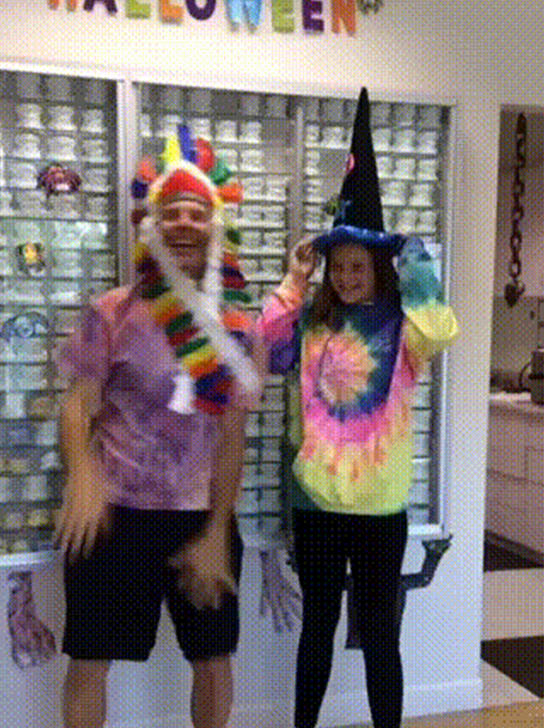 Two children in Halloween costumes pose indoors near shelving and colorful decorations.