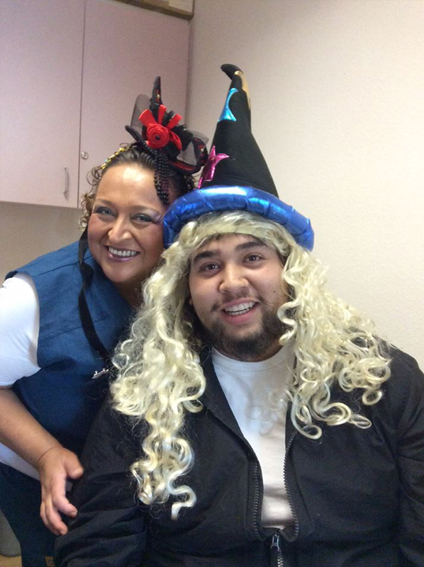 Two people wearing festive blue wizard hats with blonde wigs smile together indoors at an orthodontics office.