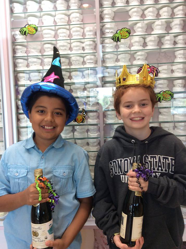 Two children wearing party hats hold trophies in front of shelves at an orthodontics office.