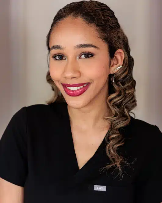 Professional headshot of woman smiling at camera wearing black scrubs in clinical setting.